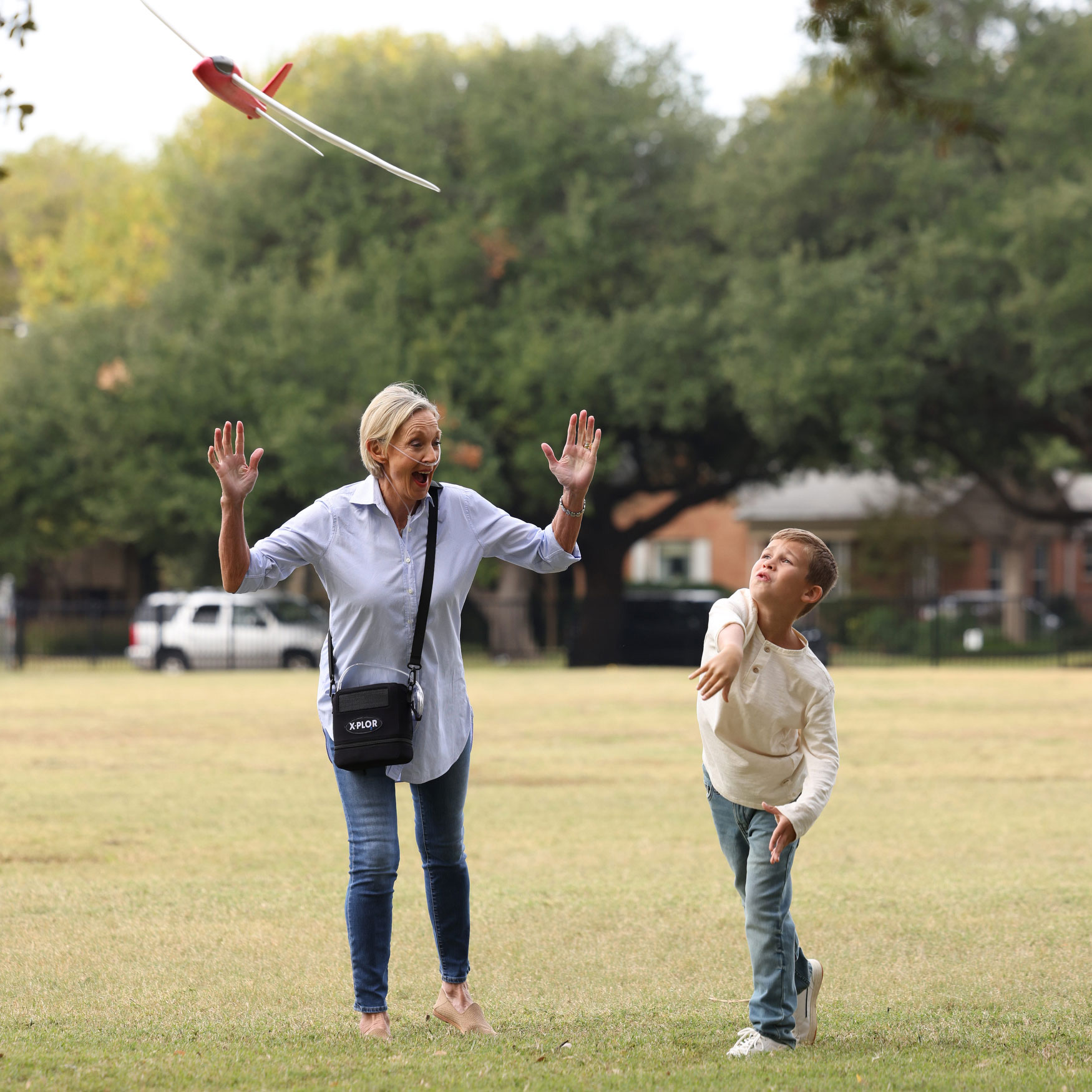 photograph of a woman using the X-PLOR portable oxygen concentrator, in a park setting, cloudy day, with young boy playing with a toy airplane