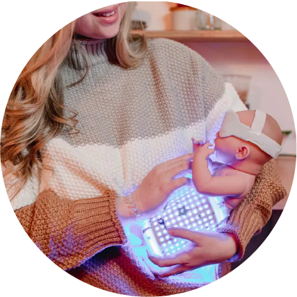 photograph of a woman holding an infant doll, with Motif BiliTouch phototherapy blanket wrapped around the doll and glowing, indicating the power is on