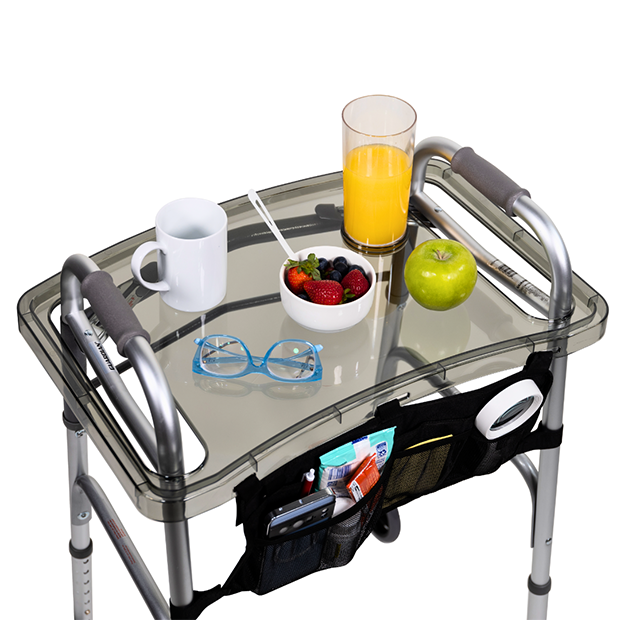 photograph of a clear acrylic tray fit over the handles of an aluminum assistive walker. the tray is holding breakfast items: a coffee cup, a glass of orange juice, a bowl of food, and an apple. a folded pair of glasses sits next to the coffee cup.