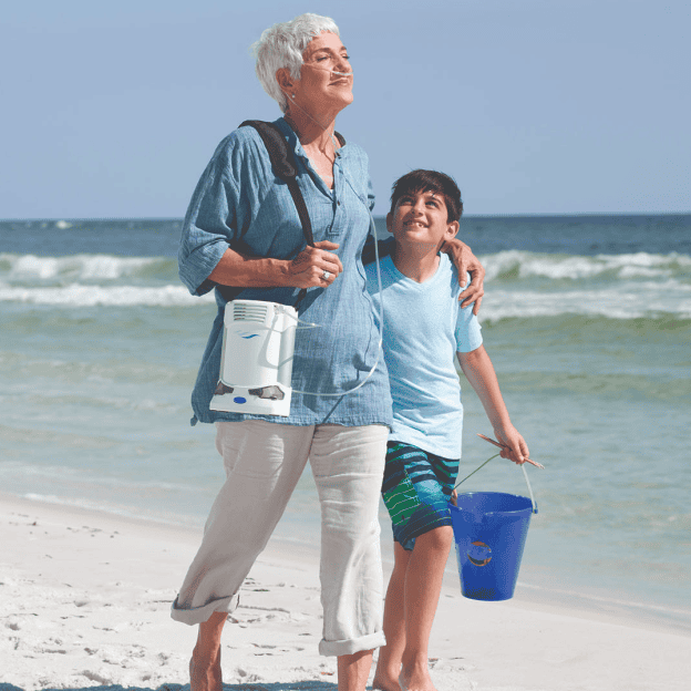 Woman and young boy walking on a beach on a sunny day with woman holding the CAIRE FreeStyle Comfort Portable Oxygen Concentrator