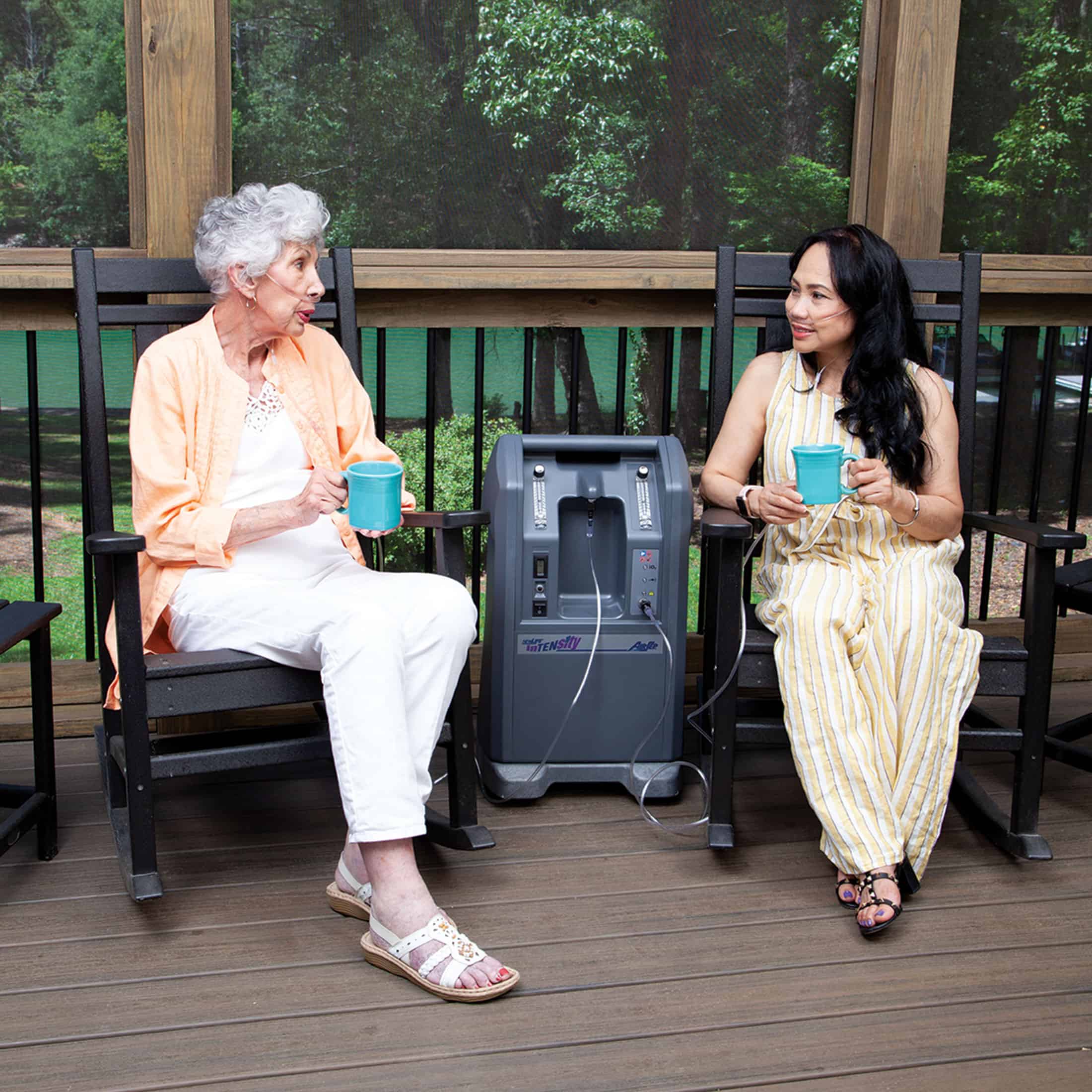 Older woman and younger woman seated on a porch simultaneously sharing oxygen flow from a single CAIRE NewLife Intensity oxygen concentrator